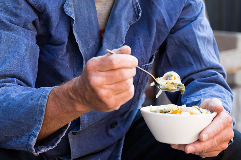 Een man met een blauw overal aan eet kippensoep